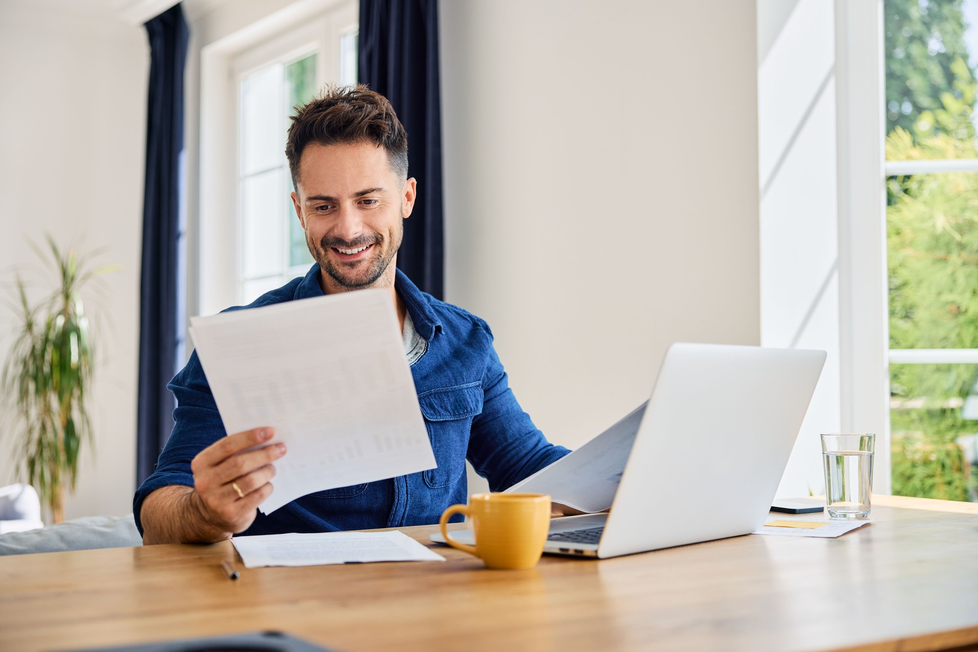 Homme heureux regardant des documents financiers à la maison assis à une table avec ordinateur portable dans le salon